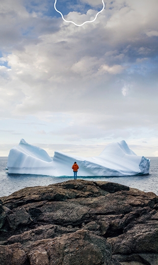 Person in an orange jacket stands on a rocky cliff, facing a massive iceberg in the ocean.