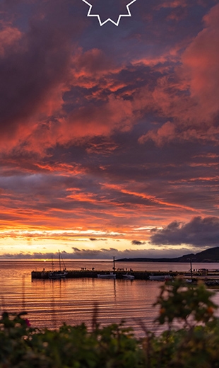 Sunset over a calm lake with a small pier, silhouetted against vibrant orange and purple clouds.